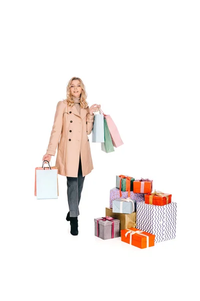 Belle jeune femme tenant des sacs à provisions et regardant la caméra tout en se tenant près de boîtes-cadeaux isolées sur blanc — Photo de stock