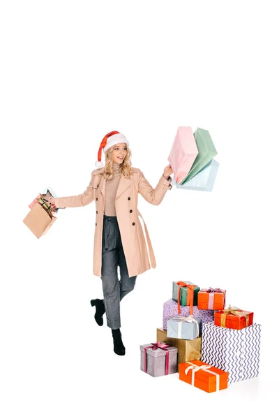 Joyeuse jeune femme en santa chapeau tenant des sacs à provisions et marchant près de cadeaux de Noël isolé sur blanc — Photo de stock