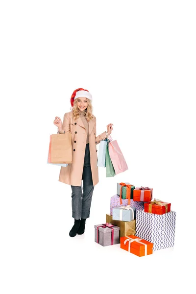 Schöne junge Frau mit Weihnachtsmannmütze, Einkaufstaschen in der Hand und lächelnd in die Kamera, während sie in der Nähe von Geschenkboxen isoliert auf weiß steht — Stockfoto