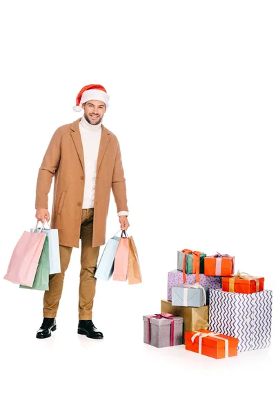 Schöner junger Mann mit Weihnachtsmannmütze, Einkaufstaschen in der Hand und lächelnd in die Kamera, während er in der Nähe von Geschenkboxen steht, isoliert auf weiß — Stockfoto