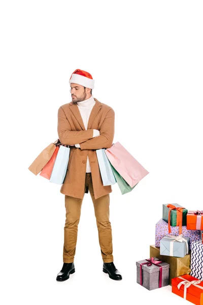 Молодий чоловік у капелюсі Санта тримає сумки, стоячи біля різдвяних подарунків ізольовані на білому — стокове фото