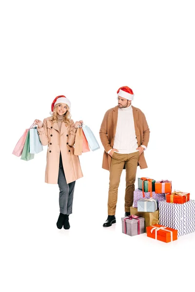 Junger Mann mit Weihnachtsmütze steht in der Nähe von Weihnachtsgeschenken und schaut lächelnde Mädchen mit Einkaufstüten auf weißem Grund an — Stockfoto