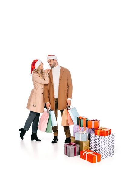 Hermosa pareja joven en sombreros de santa mirándose mientras están de pie con bolsas de compras cerca de regalos de Navidad aislados en blanco - foto de stock