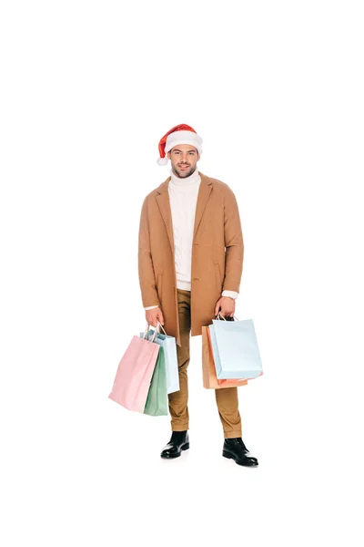 Schöner bärtiger junger Mann mit Weihnachtsmannmütze, Einkaufstüten in der Hand und in die Kamera lächelnd, isoliert auf weiß — Stockfoto