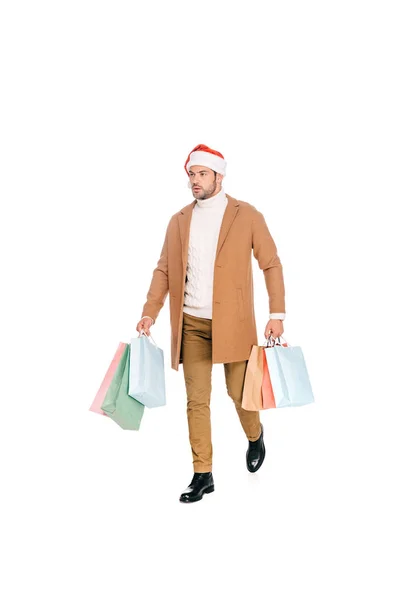 Visão comprimento total do jovem em santa chapéu segurando sacos de compras e andando isolado em branco — Fotografia de Stock