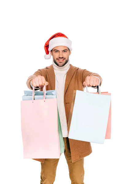 Guapo feliz joven en santa hat sosteniendo bolsas de compras y sonriendo a la cámara aislado en blanco - foto de stock