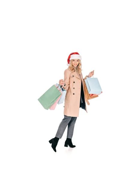 Vue pleine longueur de belle jeune femme heureuse dans le chapeau santa tenant des sacs à provisions et souriant à la caméra isolée sur blanc — Photo de stock