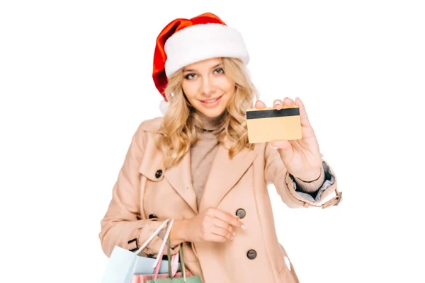 Bella sorridente giovane donna in cappello di Babbo Natale che tiene borse della spesa e carta di credito isolata su bianco — Foto stock