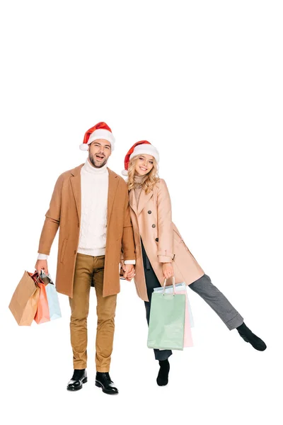 Hermosa feliz joven pareja en santa sombreros sosteniendo bolsas de compras y sonriendo a la cámara aislada en blanco - foto de stock