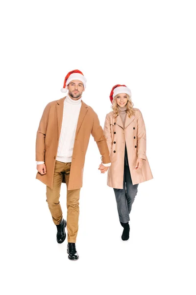 Beautiful young couple in santa hats holding hands and smiling at camera isolated on white — Stock Photo