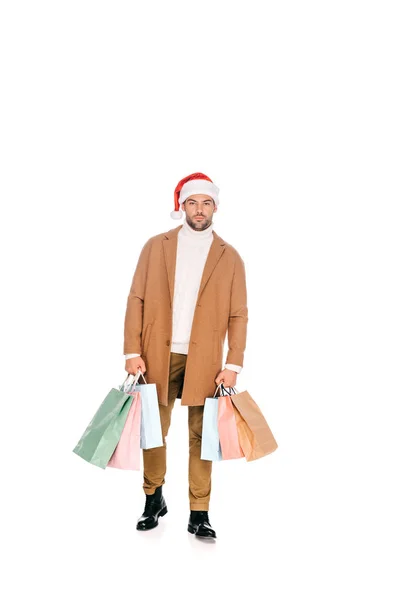 Vue pleine longueur de beau jeune homme en santa chapeau tenant des sacs à provisions et regardant la caméra isolée sur blanc — Photo de stock