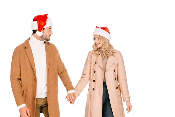 Beautiful young couple in santa hats holding hands and looking at each other isolated on white — Stock Photo
