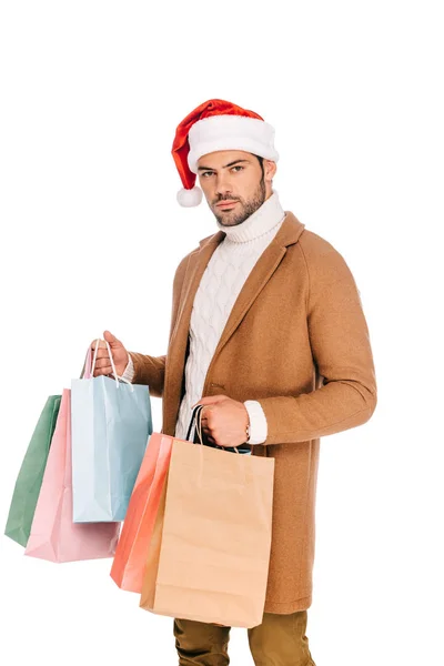 Handsome young man in santa hat holding shopping bags and looking at camera isolated on white — Stock Photo