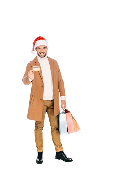 Sorridente giovane in cappello di Babbo Natale in possesso di carta di credito e borse della spesa isolate su bianco — Foto stock