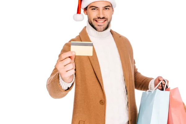 Felice giovane uomo in cappello di Babbo Natale in possesso di carta di credito e borse della spesa isolato su bianco — Foto stock