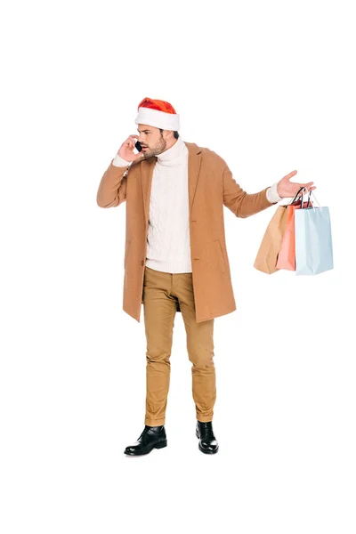 Emotional young man in santa hat holding shopping bags and talking by smartphone isolated on white — Stock Photo