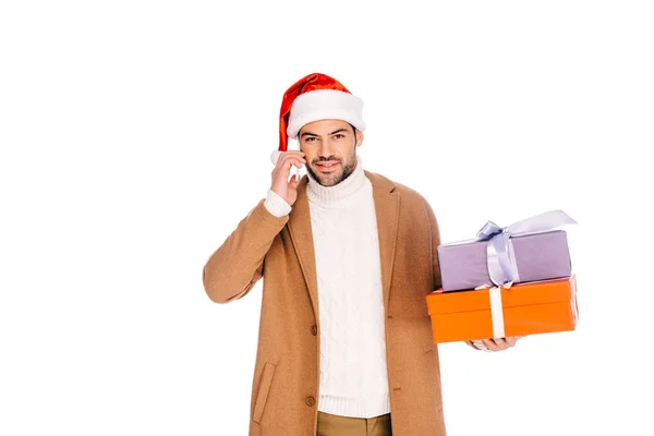 Joven en santa hat sosteniendo cajas de regalo y hablando por teléfono inteligente aislado en blanco - foto de stock