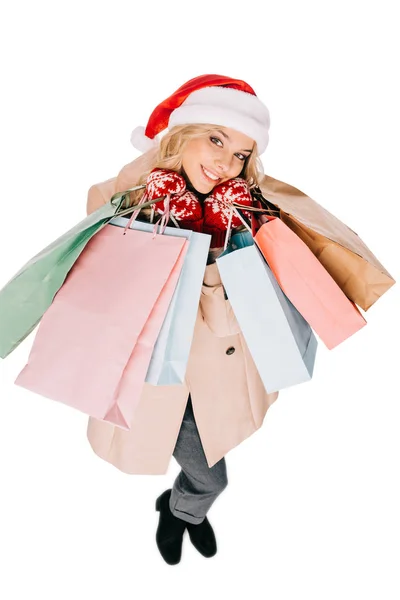 Vista ad alto angolo di felice giovane donna in cappello di Babbo Natale e guanti che tengono borse della spesa e sorridono alla fotocamera isolata su bianco — Foto stock