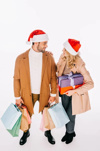 Vista de ángulo alto de pareja joven en sombreros de santa celebración de regalos y bolsas de compras aislados en blanco - foto de stock