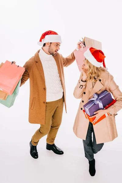 Vista de ángulo alto de pareja joven en sombreros de santa celebración de cajas de regalo y bolsas de compras aisladas en blanco - foto de stock