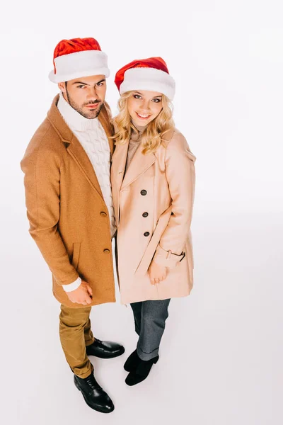 Vista de ángulo alto de hermosa pareja joven en sombreros de santa sonriendo a la cámara aislada en blanco - foto de stock