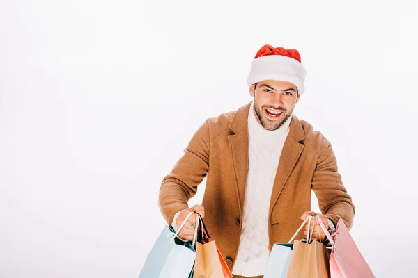 Glücklicher junger Mann mit Weihnachtsmannmütze, Einkaufstüten in der Hand und lächelnd in die Kamera, isoliert auf weiß — Stockfoto