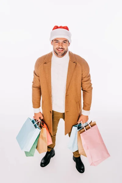 Vista de ángulo alto de hombre joven feliz en sombrero de santa celebración de bolsas de compras y sonriendo a la cámara aislada en blanco - foto de stock