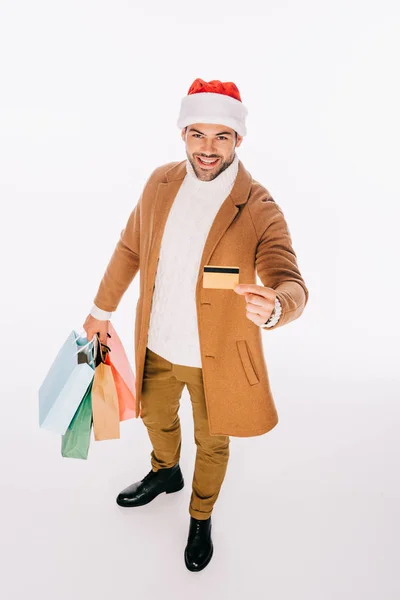 Alto angolo di vista del giovane sorridente in cappello di Babbo Natale in possesso di carta di credito e borse della spesa isolate su bianco — Foto stock