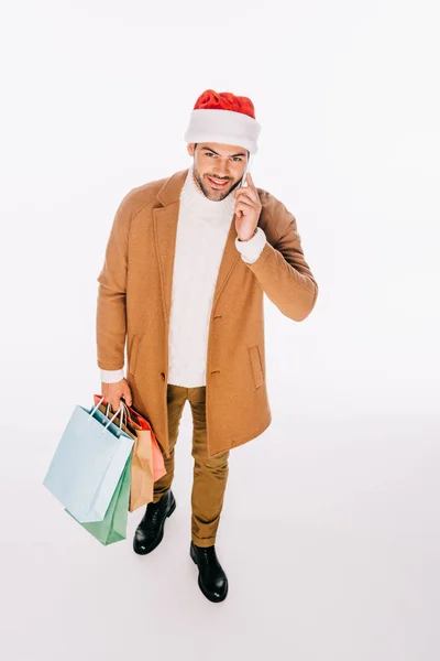 Vista ad alto angolo di giovane uomo sorridente che tiene borse della spesa e parla da smartphone isolato su bianco — Foto stock