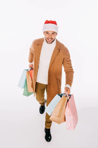 Vista ad alto angolo di bel giovane uomo in cappello di Babbo Natale che tiene borse della spesa e sorride alla fotocamera isolata su bianco — Foto stock