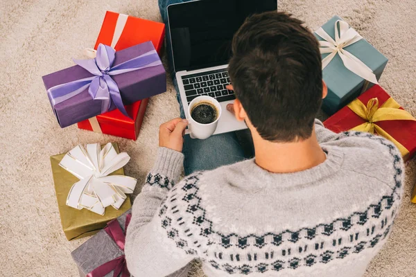 High angle view of man with cup of coffee sitting between gift boxes and using laptop with blank screen — Stock Photo