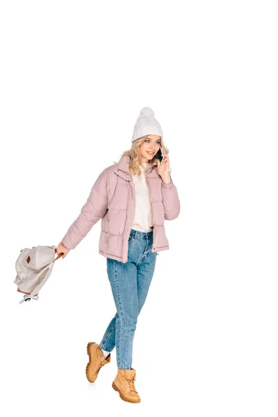 Hermosa mujer joven sonriente sosteniendo la mochila y hablando por teléfono inteligente aislado en blanco - foto de stock