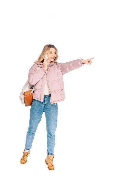 Smiling young woman with backpack talking by smartphone and pointing away with finger isolated on white — Stock Photo