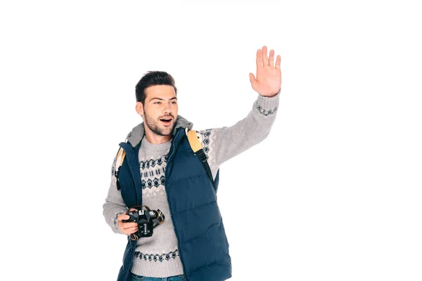 Усміхнений молодий чоловік з рюкзаком тримає камеру і махає рукою ізольовано на білому — стокове фото