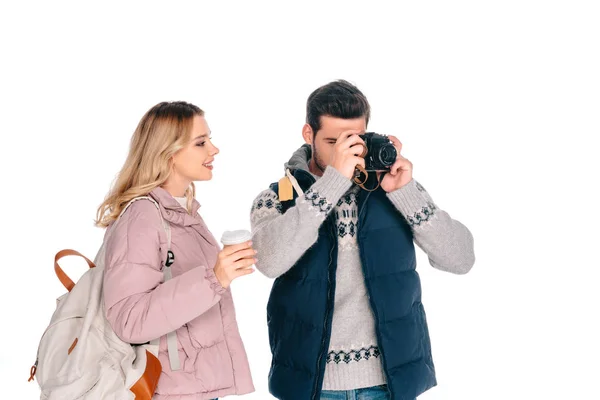 Sonriente joven con mochila sosteniendo una taza de papel y mirando a un hombre guapo fotografiando con cámara aislada en blanco - foto de stock