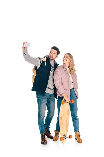 Усміхнена молода пара з рюкзаком і скейтбордом бере селфі зі смартфоном ізольовано на білому — стокове фото