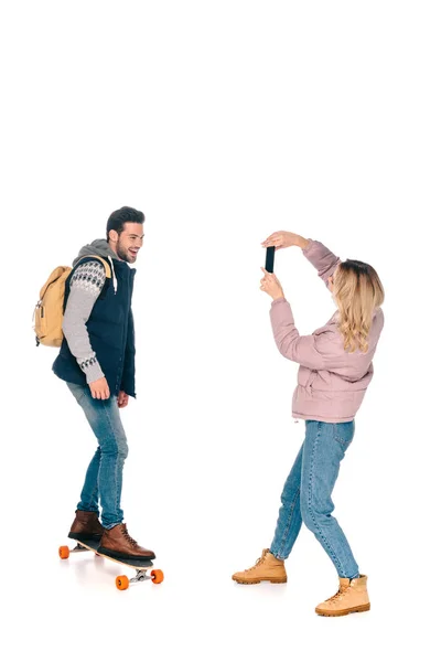 Молода жінка з смартфоном фотографує усміхненого чоловіка з рюкзаком їзда дошка — стокове фото