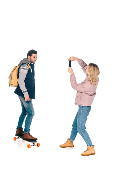 Young woman with smartphone photographing handsome man with backpack riding longboard — Stock Photo