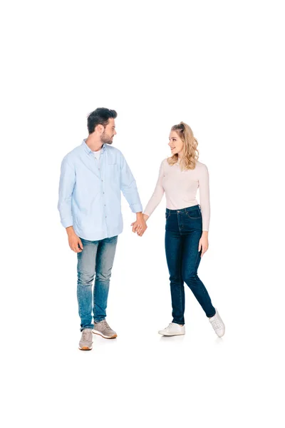 Вид в полный рост счастливой молодой пары, держащейся за руки и улыбающейся друг другу на белом фоне — стоковое фото