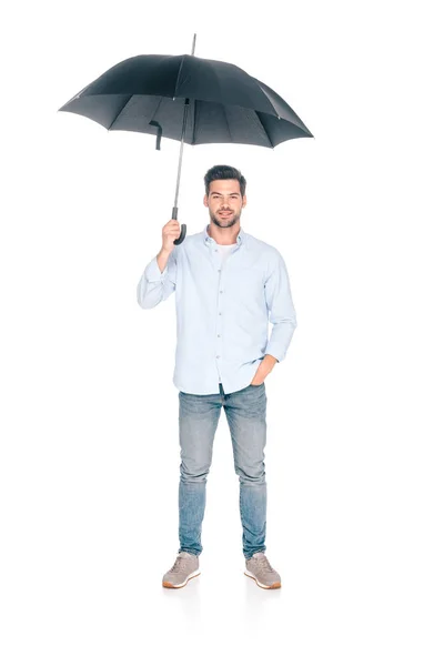 Vista completa de joven guapo sosteniendo paraguas negro y mirando a la cámara aislada en blanco - foto de stock