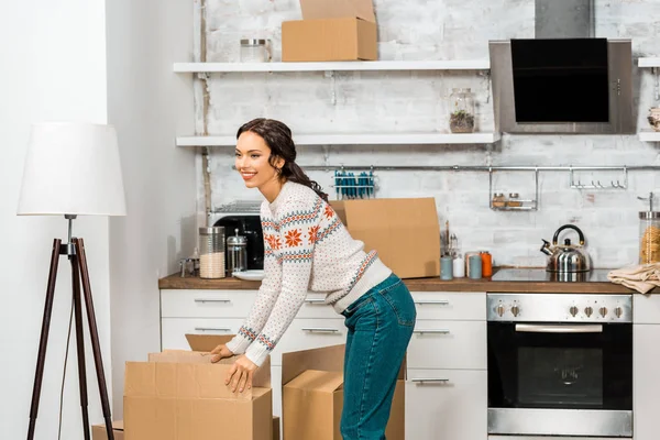 Attractive young woman standing near cardboard boxes in kitchen at new home — Stock Photo