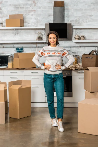 Jovem mulher de pé com as mãos na cintura perto de caixas de papelão na cozinha em nova casa — Fotografia de Stock