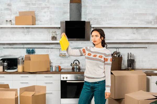 Jovem mulher segurando sujo pano amarelo na cozinha com caixas de papelão em nova casa — Fotografia de Stock