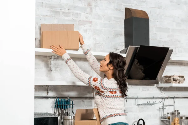Messa a fuoco selettiva di giovane donna che prende scatola di cartone dallo scaffale in cucina a nuova casa — Foto stock