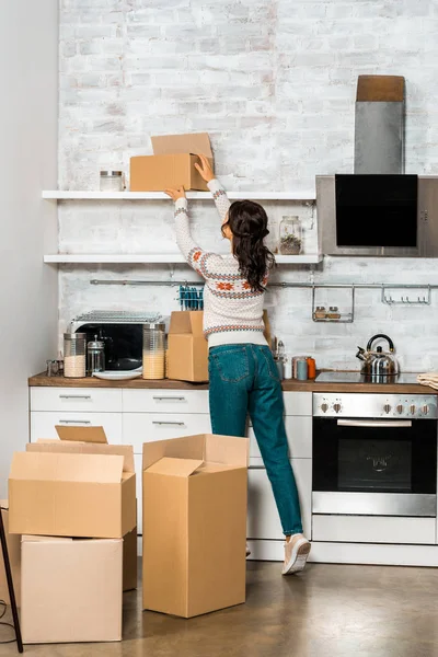 Вид сзади молодая женщина берет картонную коробку с полки на кухне в новом доме — стоковое фото