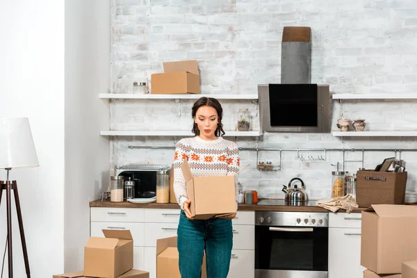 Удивленная молодая женщина смотрит в картонную коробку на кухне нового дома — стоковое фото