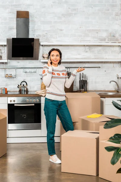 Attractive young woman talking on smartphone and doing shrug gesture during relocation in new home — Stock Photo