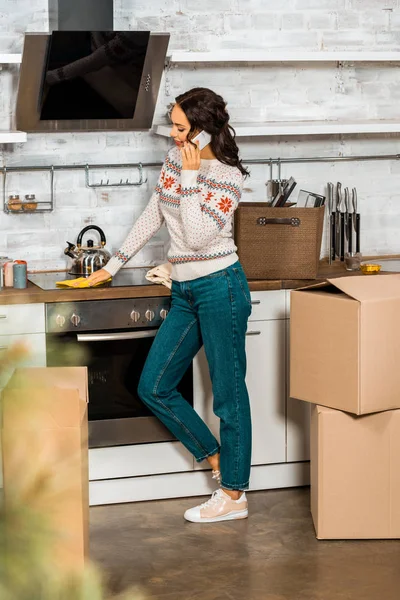 Side view of attractive woman talking on smartphone in kitchen with cardboard boxes during relocation in new home — Stock Photo