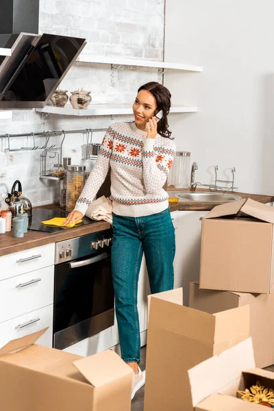 Young attractive woman talking on smartphone in kitchen with cardboard boxes during relocation in new home — Stock Photo