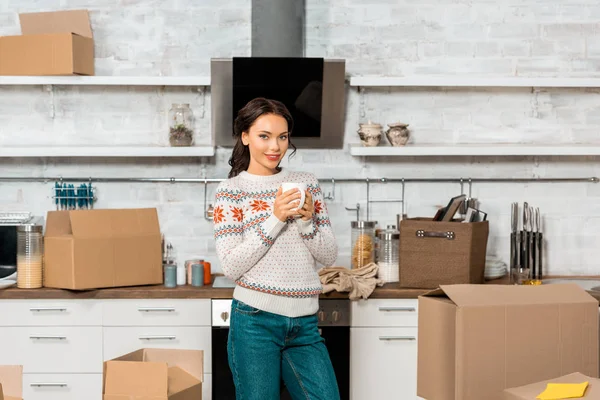 Selective focus of beautiful woman holding cup of coffee in kitchen with cardboard boxes during relocation in new home — Stock Photo